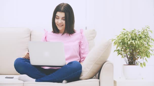 Schattig Kaukasisch brunette meisje zitten op de bank in lotos positie, met laptop, surfen op het internet, zuchten, op zoek teleurgesteld. — Stockvideo