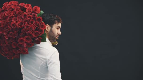 Όμορφος άντρας με μεγάλο μπουκέτο τριαντάφυλλα στον ώμο να στέκεται στην πλάτη του στην κάμερα, να γυρίζει, να χαμογελάει, να σηκώνει τα φρύδια, να κοιτάζει αποπλανητικά. — Αρχείο Βίντεο