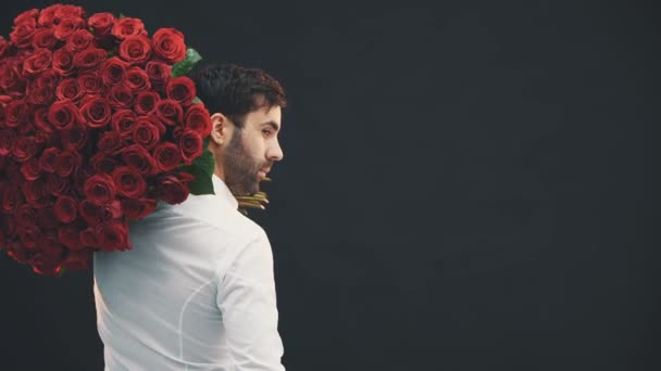 Hombre guapo con gran ramo de rosas en el hombro de pie la espalda a la cámara, girando, sonriendo, levantando cejas, mirando seductor . — Vídeos de Stock