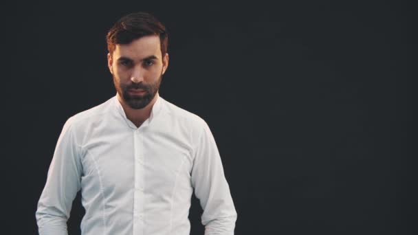 Serious young man in white shirt, standing on the black background, looking confidently at the camera. — Stock Video