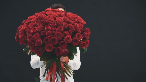 Verleidelijke man staand, met een groot boeket rode rozen, wenkbrauwen omhoog, glimlachend, verstopt achter de bloemen. — Stockvideo