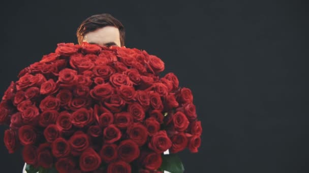 Enticing man standing, appearing from behind the large bouquet of red roses, raising his eyebrows, nodding. — Stock Video