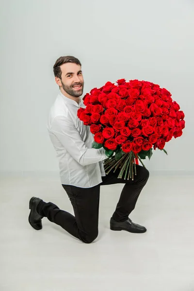 全长的照片，年轻英俊男子拿着玫瑰，看着相机，跪着时微笑着，背对着白色的背景. — 图库照片