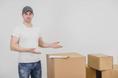 Gülümseyen genç teslimatçı, bir yığın karton paketin yanında duruyor, metin veya ürün için ellerini kopyalama alanına doğrultuyor..