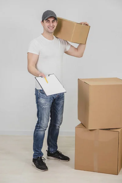 Jonge bezorger met kartonnen doos op zijn schouder, naar de camera kijkend, glimlachend, een klembord uitschuivend voor handtekening.. — Stockfoto