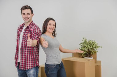 Mutlu adam ve kadın yeni dairelerinde kutular yığınının yanında duruyorlar, başparmaklarını kaldırıyorlar, gülümsüyorlar..