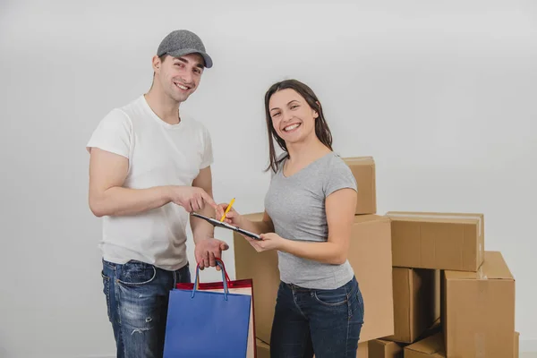 Handsome courier delivered presents in colorful bags for young woman on the occasion of her moving into new apartment. Woman is really happy and is signing the declaration. — Stock Photo, Image