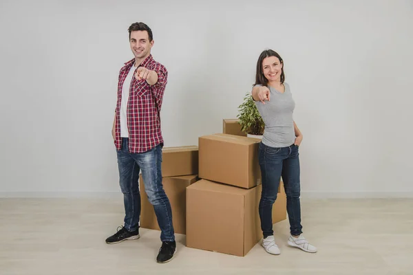 Homem e mulher animados de pé lado a lado perto da pilha de caixas de papelão em seu novo apartamento, apontando os dedos para a câmera, sorrindo . — Fotografia de Stock