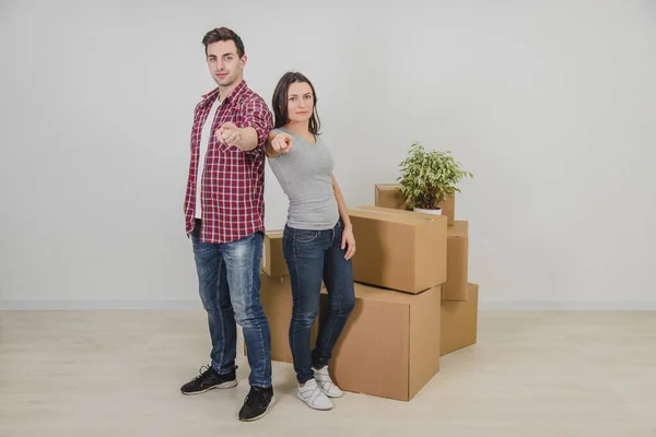 Full-length happy man and woman standing side by side near stack of carton boxes in their new apartment, pointing their fingers at the camera, smiling. — Stock Photo, Image