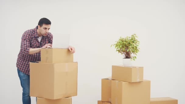 Heureux jeune homme emménagé dans une nouvelle maison. Il est debout avec un ordinateur portable sur la pile de boîtes, boire du café, à la recherche de meubles dans les boutiques en ligne, appelant pour le commander . — Video