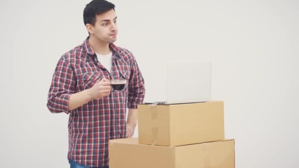 Mutlu genç adam yeni eve taşındı. Dizüstü bilgisayarla kutu yığınının üzerinde duruyor, kahve içiyor, online dükkanlarda mobilya arıyor.. — Stok video