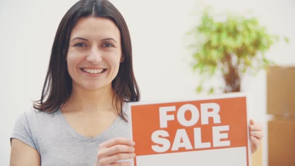 Młoda piękna kobieta trzyma talerz ze słowami na sprzedaż, oferując dom na sprzedaż i wyprowadzając się. — Wideo stockowe