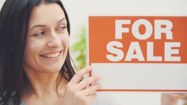 Glimlachende jonge mooie vrouw houdt een bord met woorden te koop geschreven op het, het aanbieden van huis te koop en verhuizen. — Stockvideo