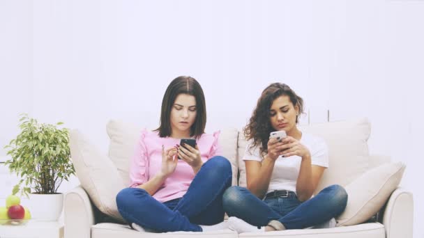 Piękne młode perwersyjne afro-amerykańskie i brunetka kaukaskie dziewczyny siedzą na kanapie w pozycji lotosu, z telefonem w rękach, wiadomości. — Wideo stockowe