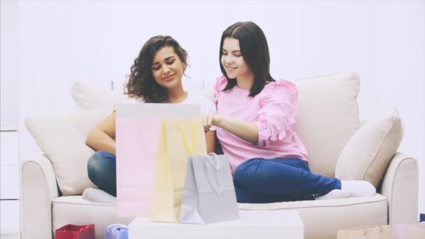 Twee vrolijke vriendinnen die thuis op de bank zitten en laptop gebruiken om online te winkelen. Wazige zakken op de voorgrond. — Stockvideo