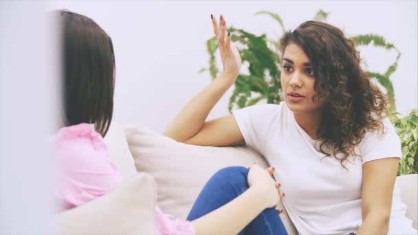 Two girl friends are talking sitting on the sofa in warm home environment. Brunette caucasian girl sitting her side to the camera, blurred. — Stock Video