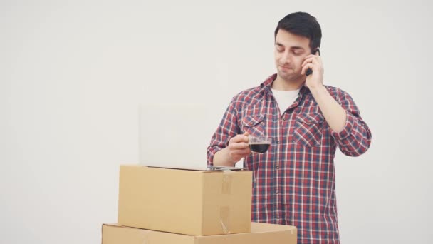 Happy young man moving into new house, standing, talking on the phone, drinking coffee, looking aroung the flat smiling. Laptop on the pile of carboard boxes. — Stock Video