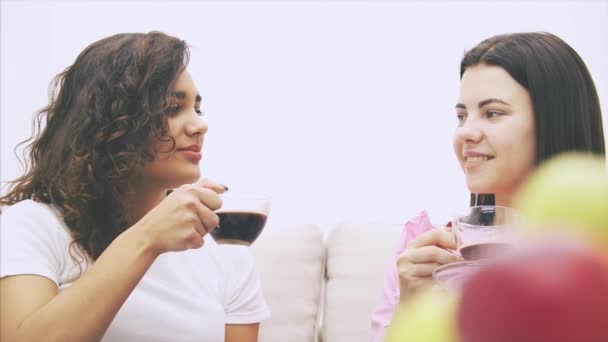 Two girl friends are talking sitting on the sofa in warm home environment, drinking coffee. Pile of apples on the blurred forefront. — Stockvideo