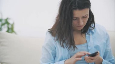 Takdire şayan bir kadın evdeki kanepede otururken akıllı telefonunu kullanmaya odaklanır. Kapatın. Uzayı kopyala 4k.