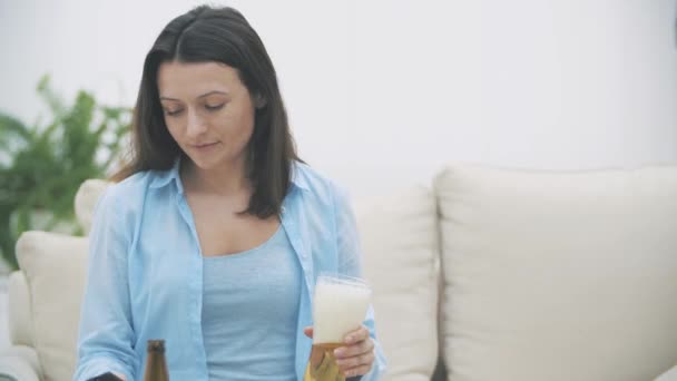Улыбающаяся и жизнерадостная женщина пьет пиво, сидит дома на диване. Принято. Закрывай. 4K . — стоковое видео
