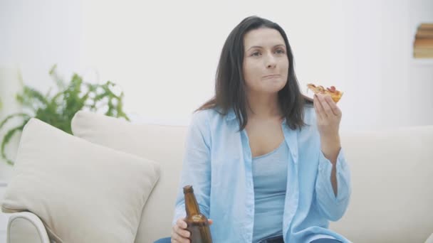 Una joven está sentada en un sofá, comiendo pizza y bebiendo cerveza. Apoya a su equipo favorito. Delicioso-delicioso. 4K . — Vídeos de Stock