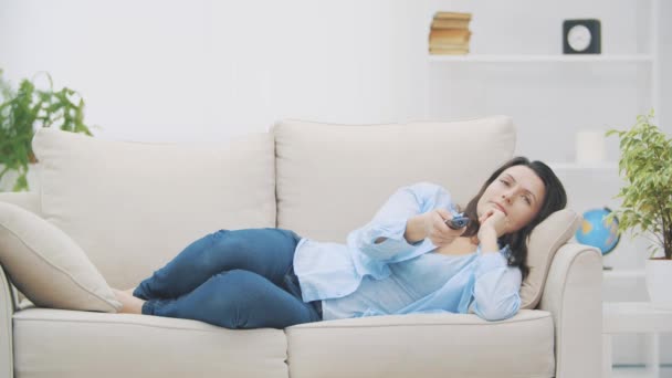 Jeune femme souriante change de chaîne avec zapper. Elle regarde la télé, allongée sur le canapé à la maison. Espace de copie. 4K. Mouvement lent . — Video