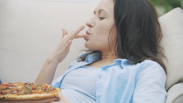 Медленно. Молодая женщина облизывает пальцы, смотрит телевизор и ест пиццу. 4K . — стоковое видео