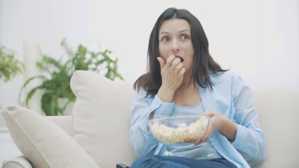 Медленно. Крупный план очаровательной женщины, которая жует поп-кукурузу, смотря телевизор с удивлением. Вкуснятина. 4K . — стоковое видео