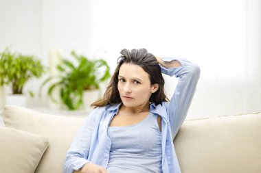 Üzgün genç bir kadın, beyaz arka planda izole bir kanepede otururken saçına dokunuyor. Olumsuz duygular gösteriyor ve saldırmaya hazır..