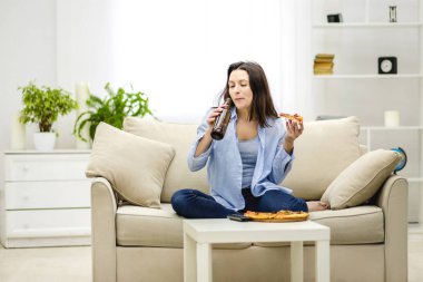 Genç ve çekici bir kadın bira içiyor ve elinde bir dilim pizza tutuyor. Bulanık ışıklı arka planda yalnız bir kadın. Boşluğu kopyala.
