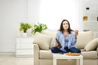Esmer kadın bir şişe bira ve bir dilim pizzacı eli tutarak çiğniyor. Bulanık ışıklı arka planda yalnız bir kadın. Boşluğu kopyala.