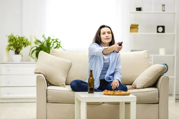 Rozkošná brunetka žena je zapínání na televizi se zapper a je připravena pohltit nevyžádané jídlo. — Stock fotografie