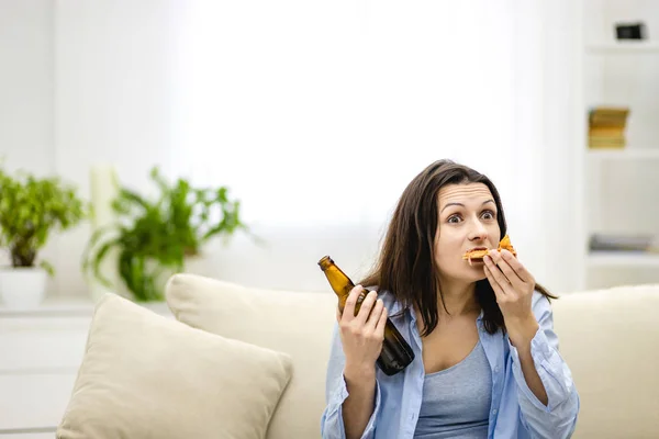 Hungry adorable woman is biting pizza and holding a bottle of beer, sitting on the sofa, full of pillows. Woman with surprised facial expression. Close up. Copy space.