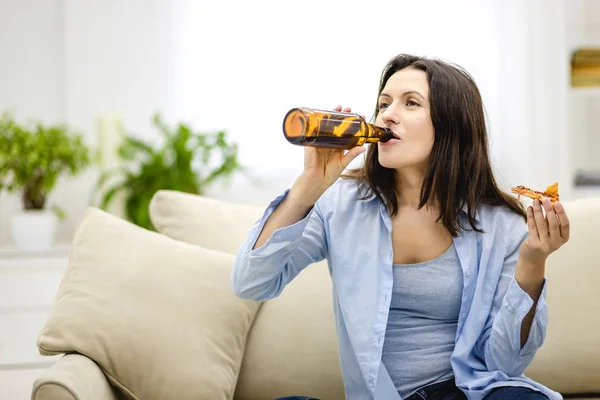 Pleased, attractive woman drinks beer and holds a slice of pizza in the other hand. Isolated woman on blurred light background. Copy space. Close up.