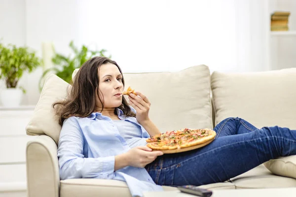 Zmęczona kobieta żuje pizzę, leżąc na białej kanapie. Ogląda programy telewizyjne, będąc na rozmytym tle.. — Zdjęcie stockowe