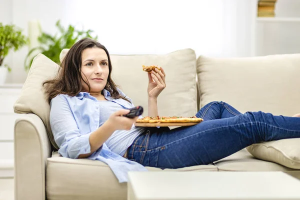 Bella donna è sdraiata sul divano a casa e sta guardando la televisione mentre mangia la pizza . — Foto Stock