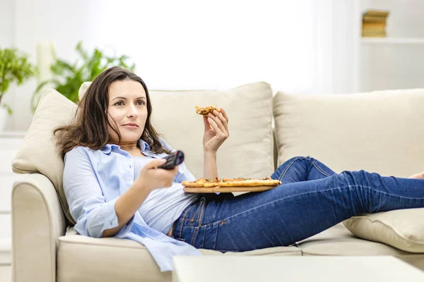 Ładna kobieta leży na kanapie w domu i ogląda telewizję jedząc pizzę.. — Zdjęcie stockowe