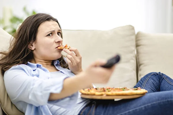 Przerażona młoda kobieta ogląda telewizję i je pizzę. Brunetka dziewczyna siedzi na kanapie z pilotem, i zmienia kanał. — Zdjęcie stockowe