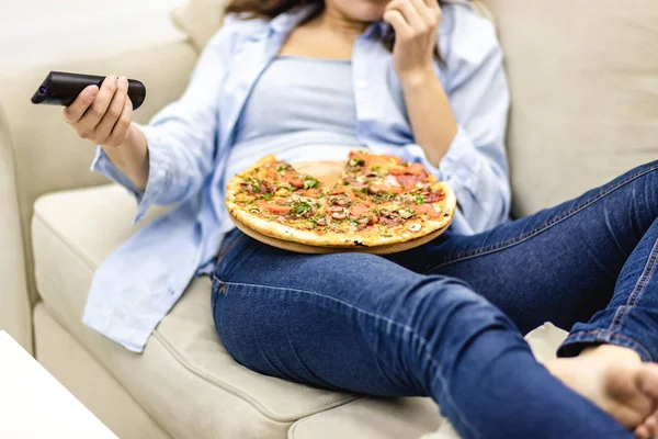 Ekin. Bacaklarını kot pantolonla kapat. Güzel bir kadın evdeki kanepede uzanıyor ve pizza yerken televizyon izliyor.. — Stok fotoğraf