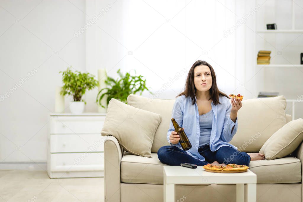 Brunette woman chews, holding a bottle of beer and a slice of pizza hands. Isolated woman on blurred light background. Copy space.