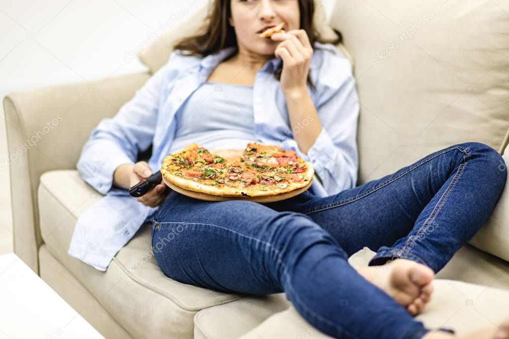 Crop. Close up legs in jeans. Pretty woman is laying on the sofa at home and is watching television while eating pizza.
