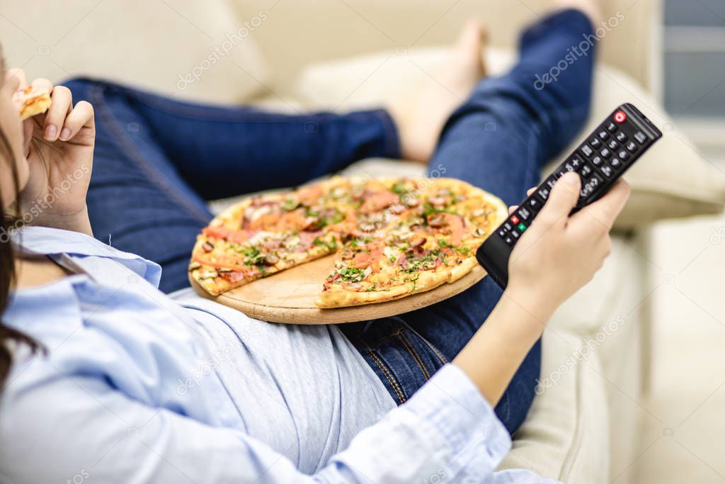 Crop. Close up legs in jeans. Unrecognizable woman is laying on the sofa at home and is watching television while eating pizza.