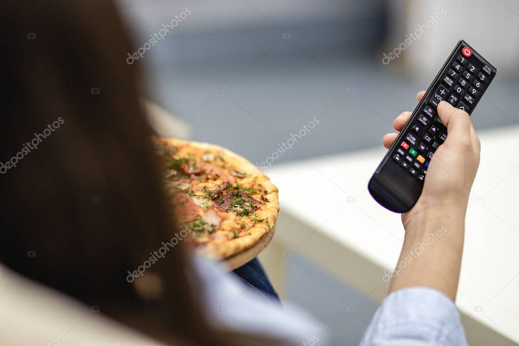 Crop. Close up remote control device. Unrecognizable woman is laying on the sofa at home and is watching television while eating pizza.