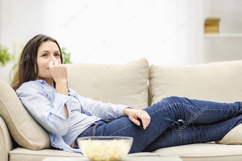 Portrait of pretty, charming, cheerful woman with straight black hair, who is eating pop corn, watching comic serial, comedy, sitting with crossed legs in living room.