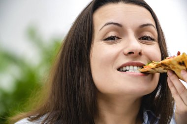 Siyah düz saçlı güzel genç bayan bir dilim sıcak pizzayı ısırıyor. O, doğrudan kameraya bakıyor. Uzayı kopyala Kapat..