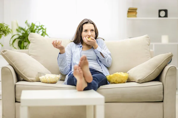 Portrét krásné, okouzlující, veselé ženy s rovnými černými vlasy, která jí popcorn a bramborové lupínky, dívá se na komiks, komedie, sedí se zkříženýma nohama v obývacím pokoji. — Stock fotografie