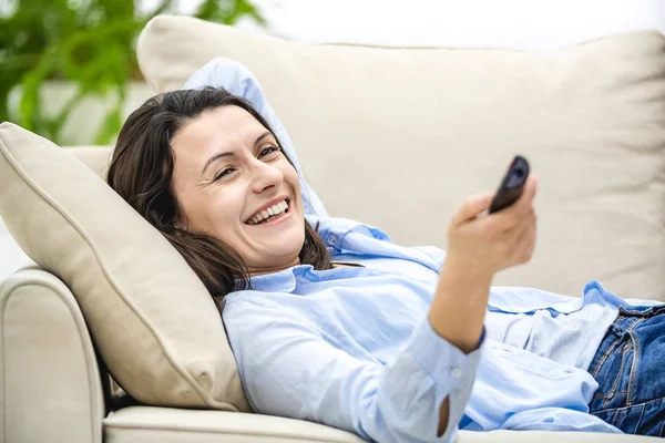 Glückliche junge Frau sitzt auf einem Sofa vor dem Fernseher. Sie lächelt breit. Ernte. — Stockfoto