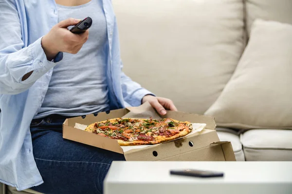 作物。 无法辨认的女人正坐在家里的沙发上，一边看电视一边吃披萨。 复制空间. — 图库照片