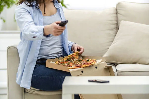 Gewas. Onherkenbare vrouw zit thuis op haar bank en kijkt televisie terwijl ze pizza eet. — Stockfoto