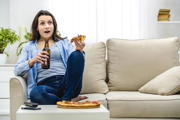 迷人而感兴趣的女人在看电视时震惊了。 吃惊的女人正坐在凌乱的沙发上吃着小吃. — 图库照片